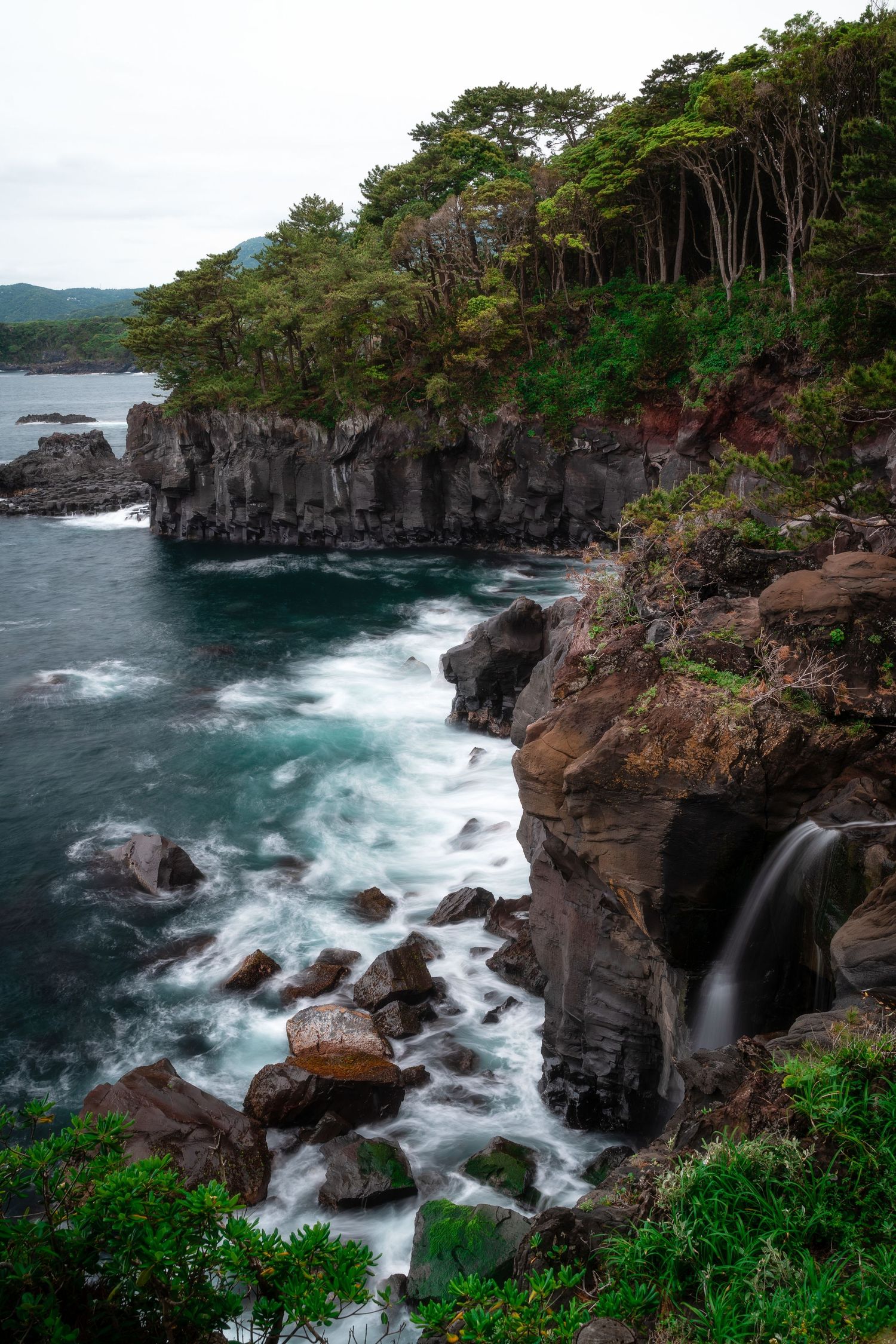 Rocky coastal cliffside with lush trees, crashing waves, and a small waterfall in nature.