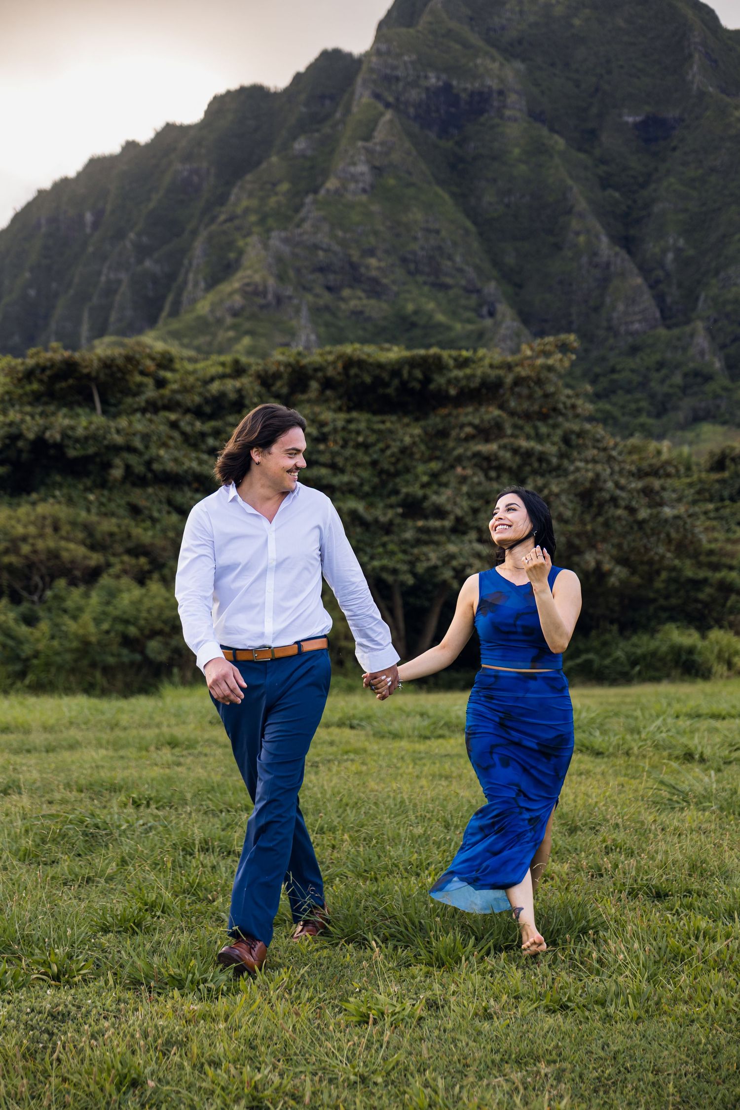 A couple holds hands and walks through a green field with dramatic mountain cliffs in the background.