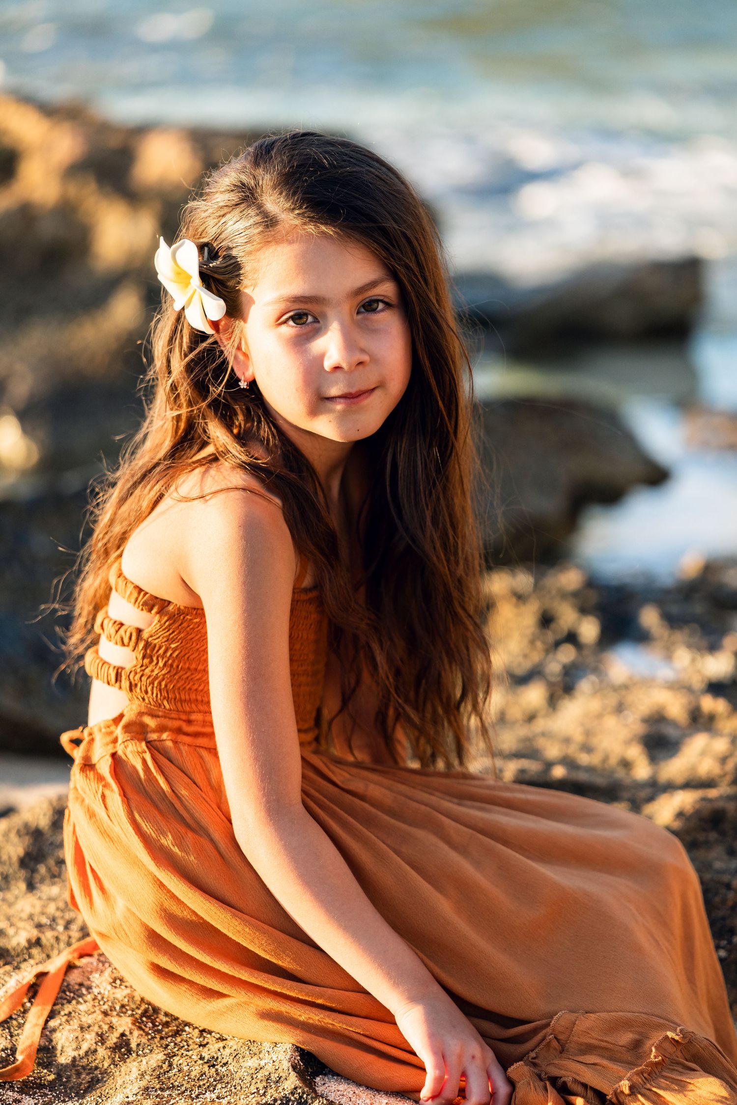 A young girl with long hair and a flower accessory sits on rocks by the ocean in a golden dress.