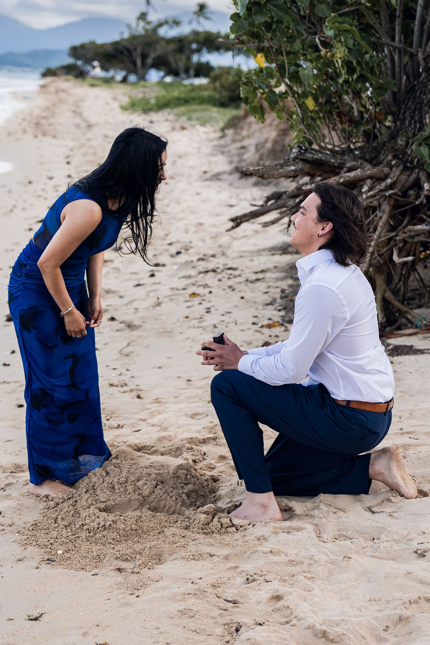 Man kneeling on sandy beach proposing to woman in blue dress at tropical shoreline.