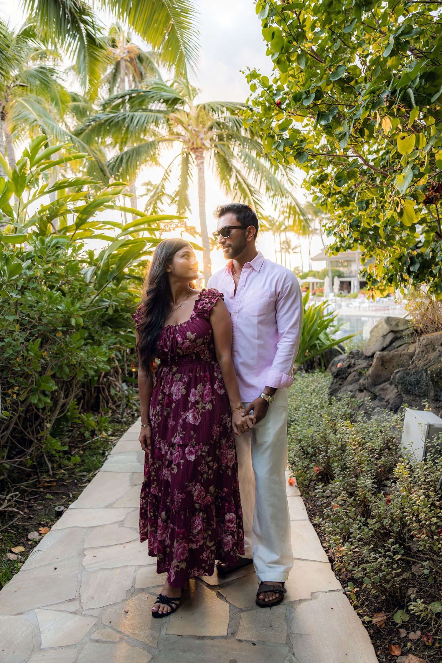 Couple holding hands on a tropical garden path, woman in floral dress, man in white shirt, lush palm trees background.