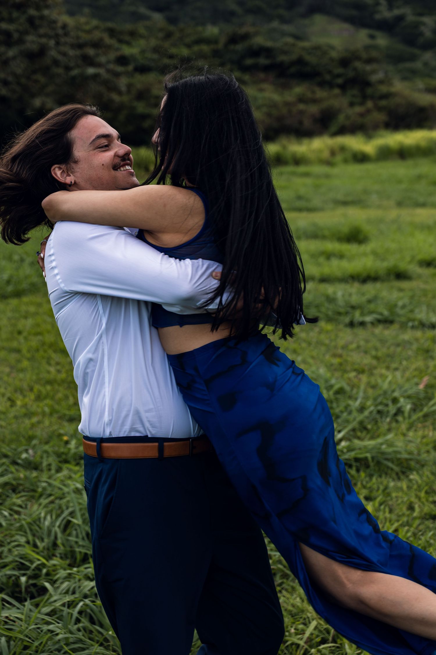 Joyful couple embracing and spinning in lush green meadow with mountains in background.