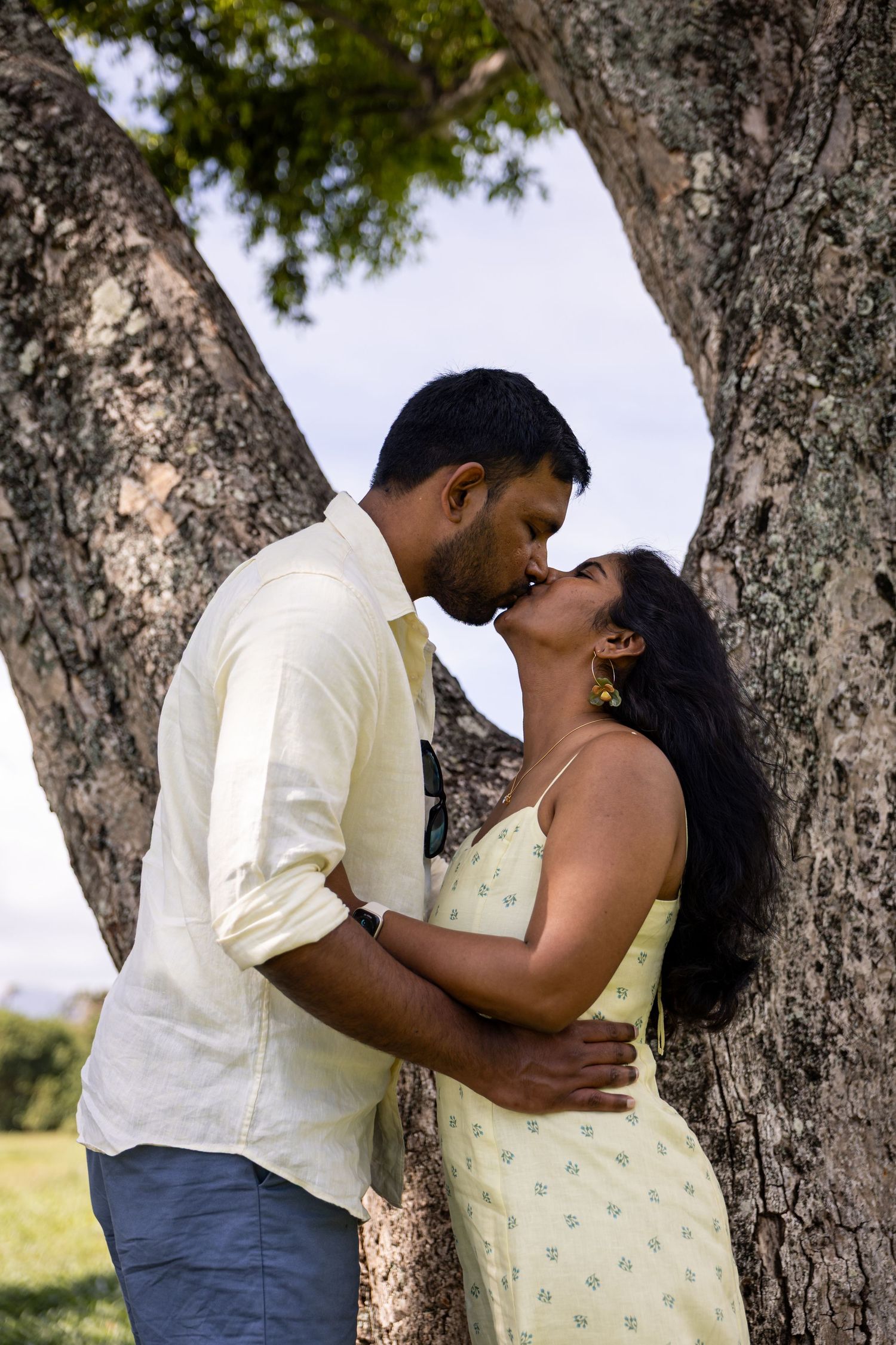 A romantic couple shares a kiss beneath a large tree outdoors, the woman in a yellow floral dress and man in white shirt.