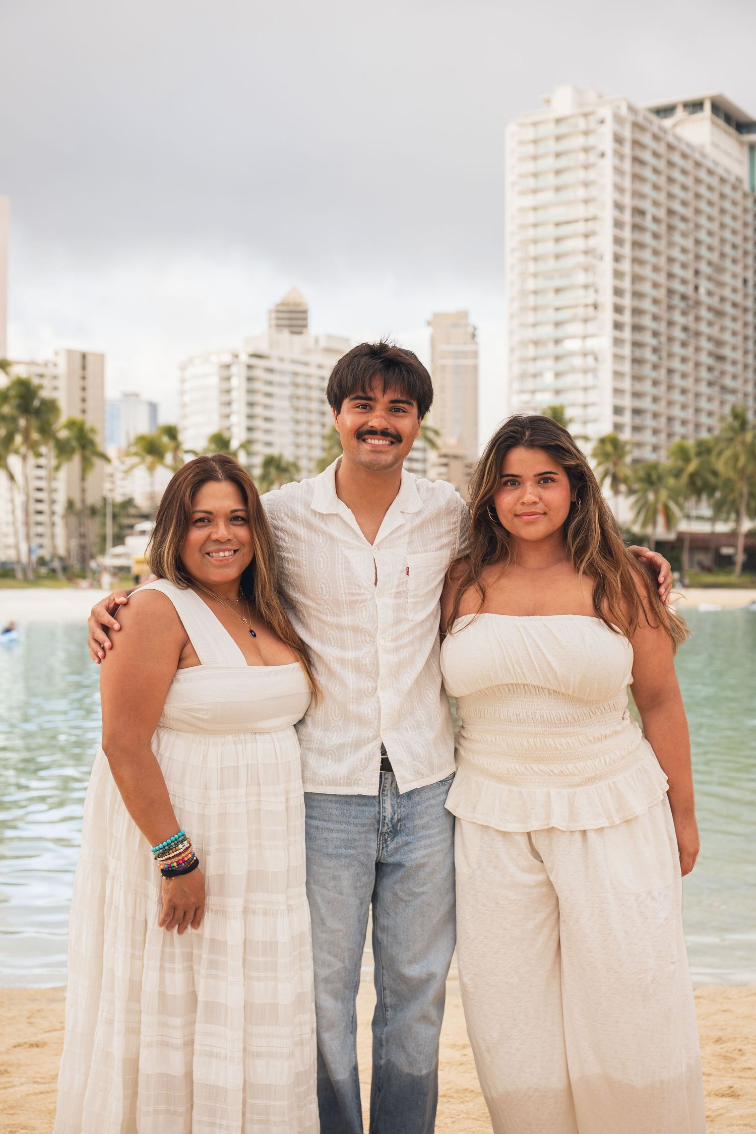 Three people in white outfits pose on a beach with palm trees and city buildings in the background.
