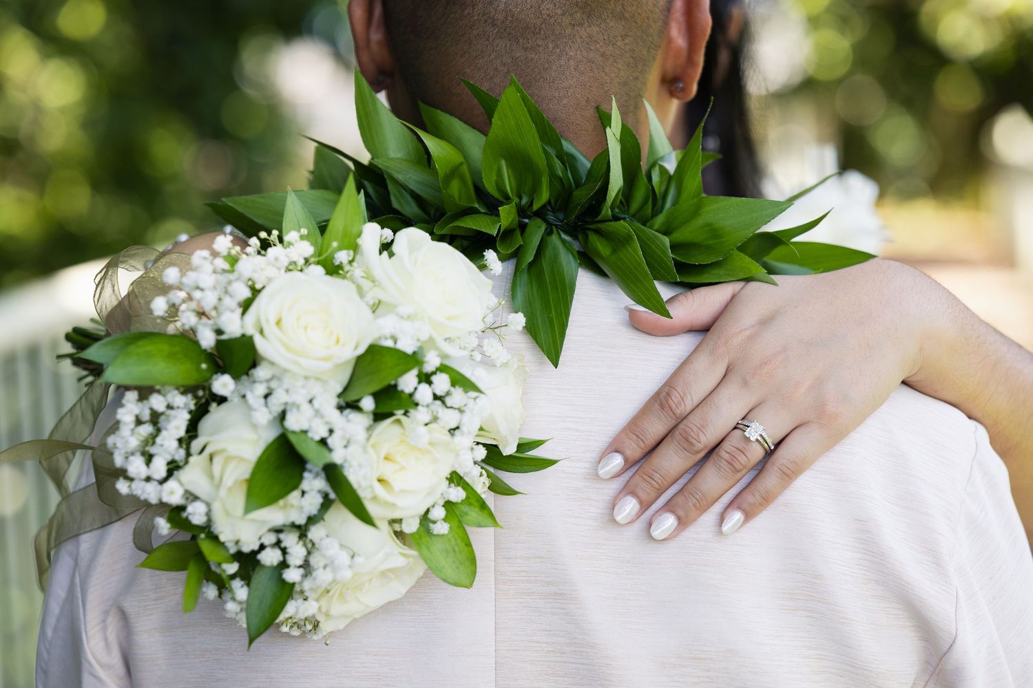 Bride holding white rose bouquet with baby's breath, resting hand on groom's shoulder, wearing diamond engagement ring.