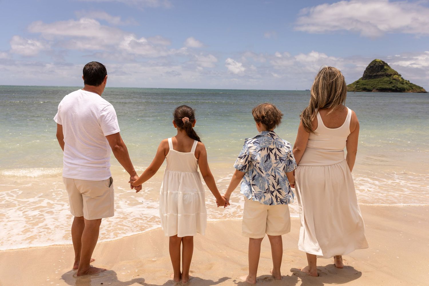 Family of four holding hands on a sunny Hawaiian beach, viewed from behind with turquoise ocean and island in background.