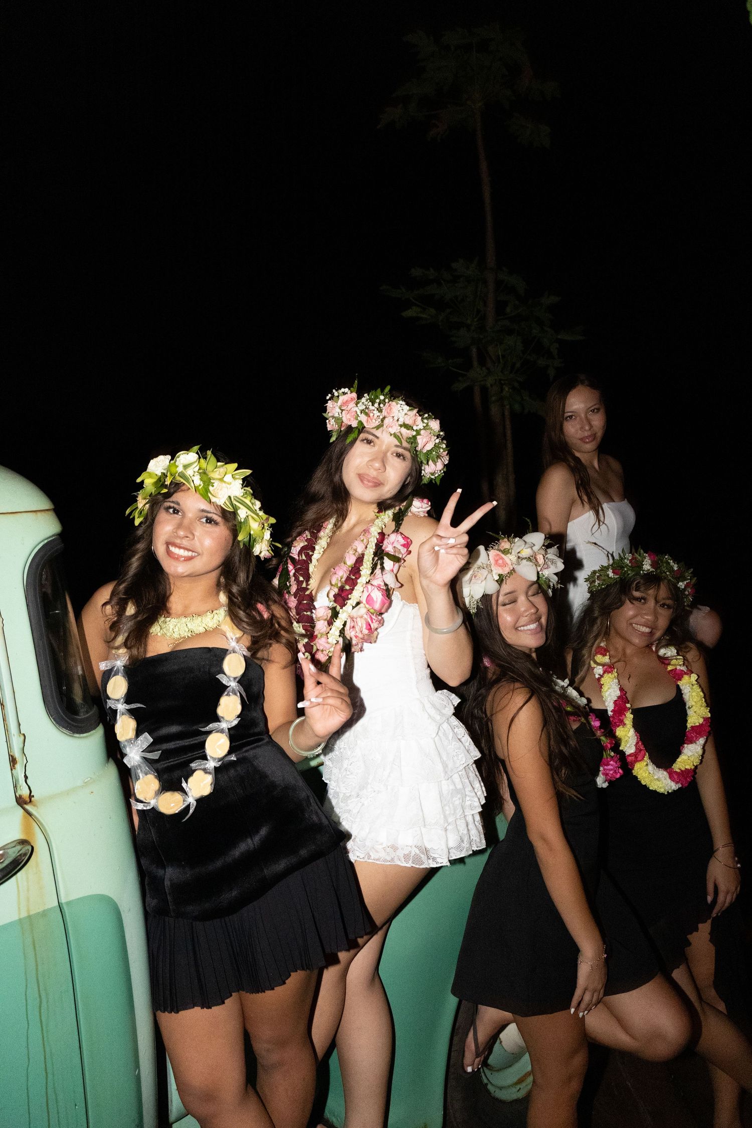 Young women in floral crowns and leis pose cheerfully at night near a vintage teal truck at a Hawaiian celebration.