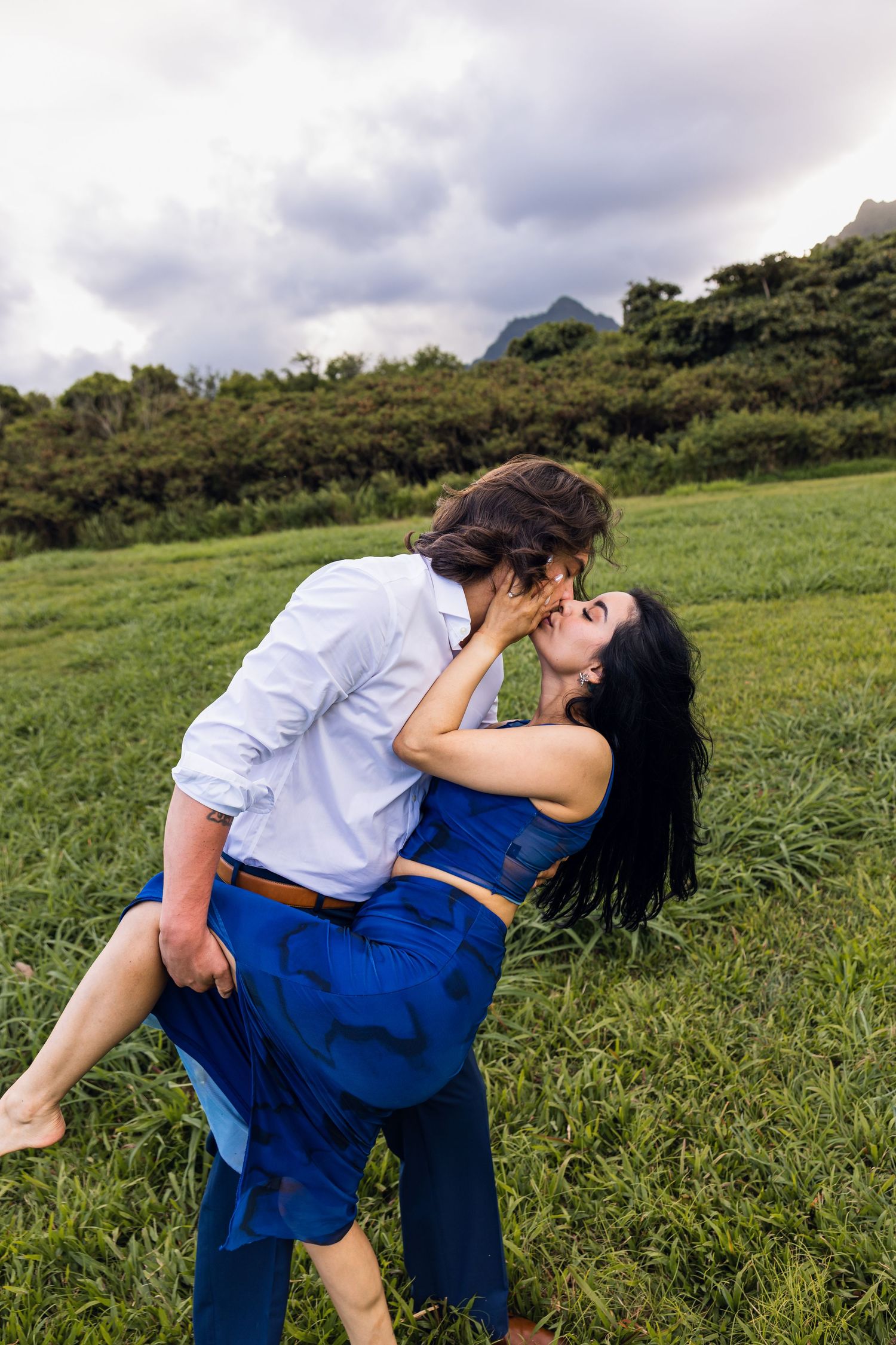 Couple sharing a romantic dip kiss in a lush green field with misty mountains in the background.
