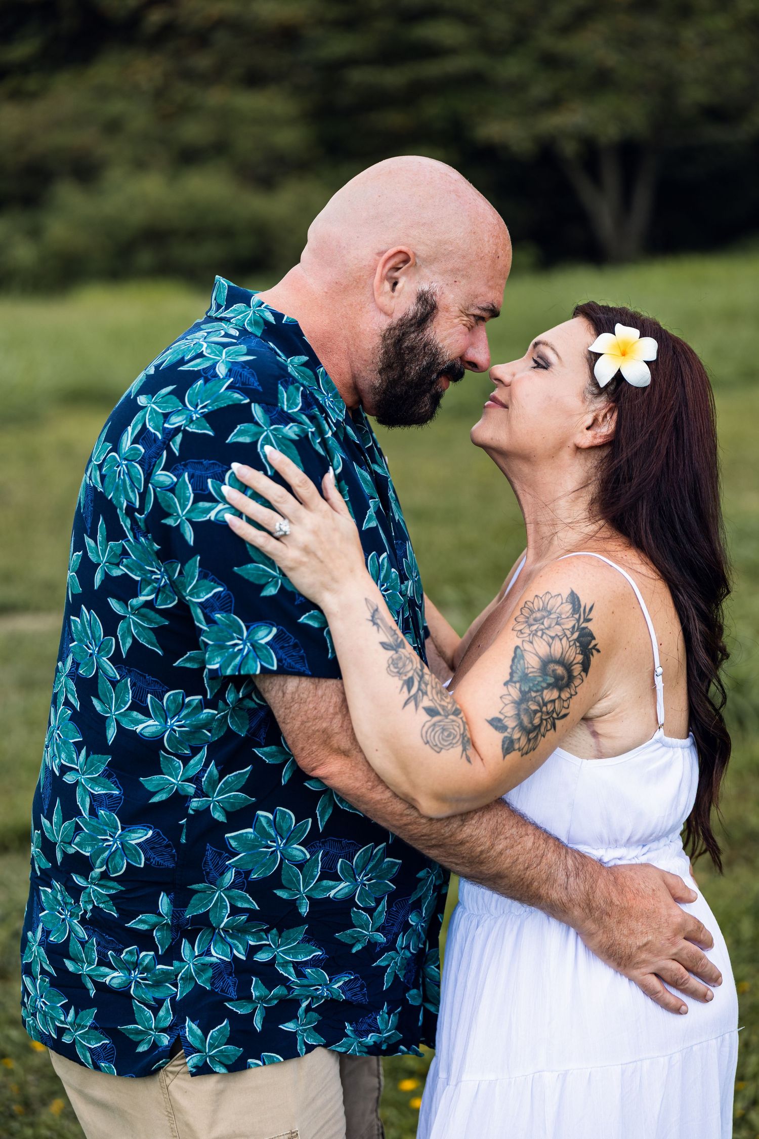 A tattooed couple in a tropical setting share a romantic moment, the woman in white dress with a plumeria flower in her hair.