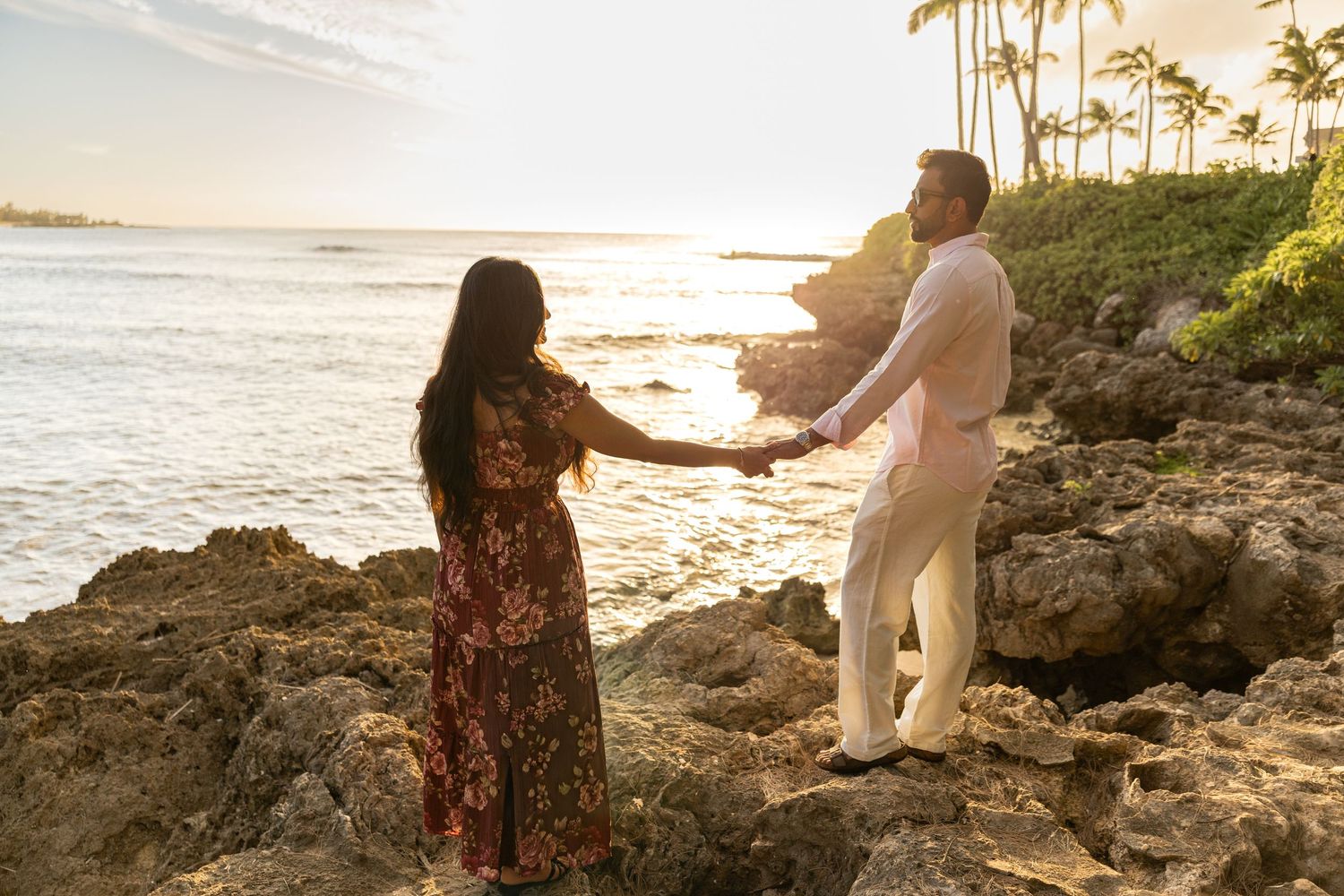 Romantic couple holding hands on rocky shoreline at sunset with palm trees and ocean in Hawaii.