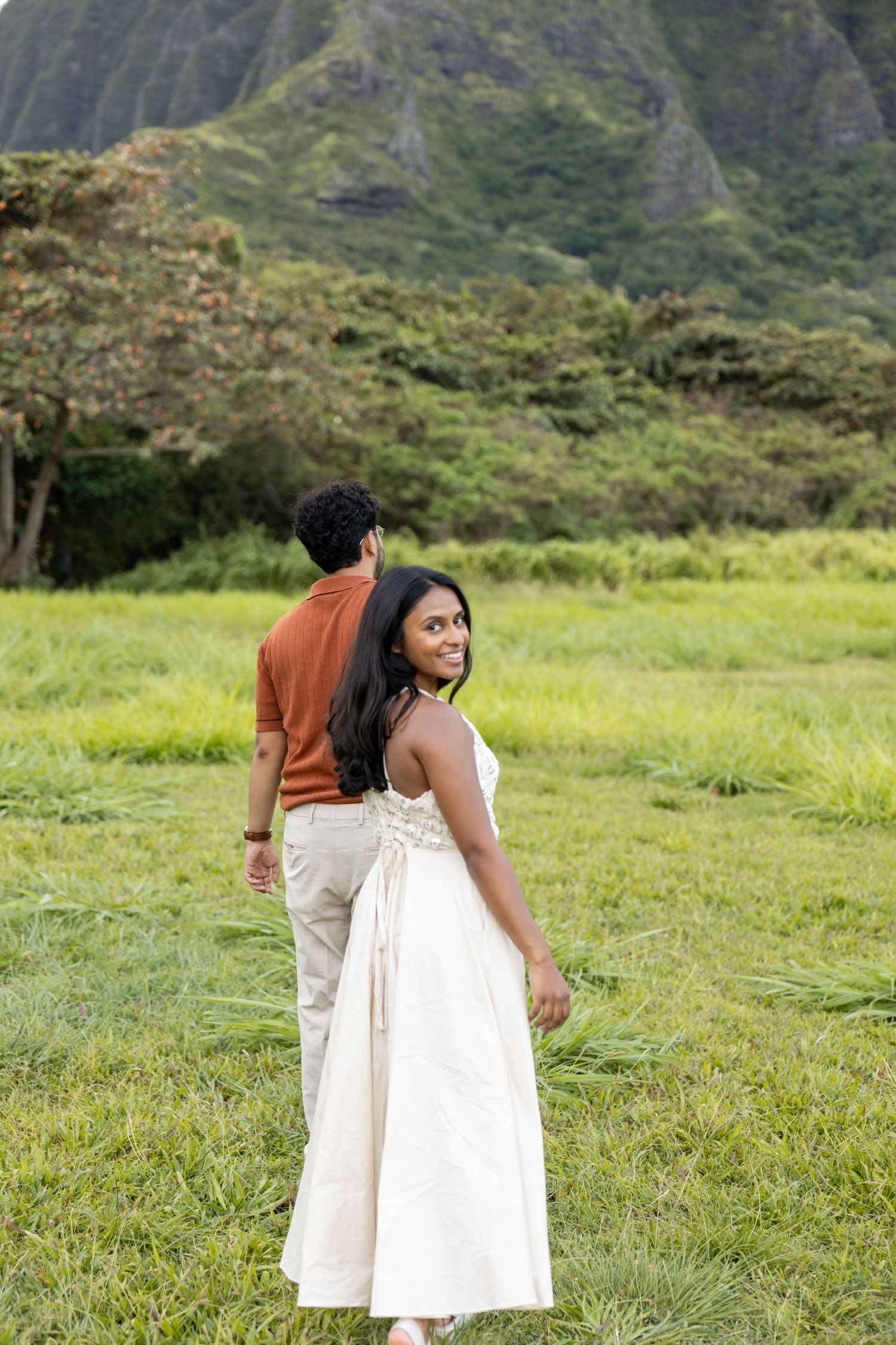 Young woman in white dress smiles over shoulder as couple walks through green Hawaiian meadow.