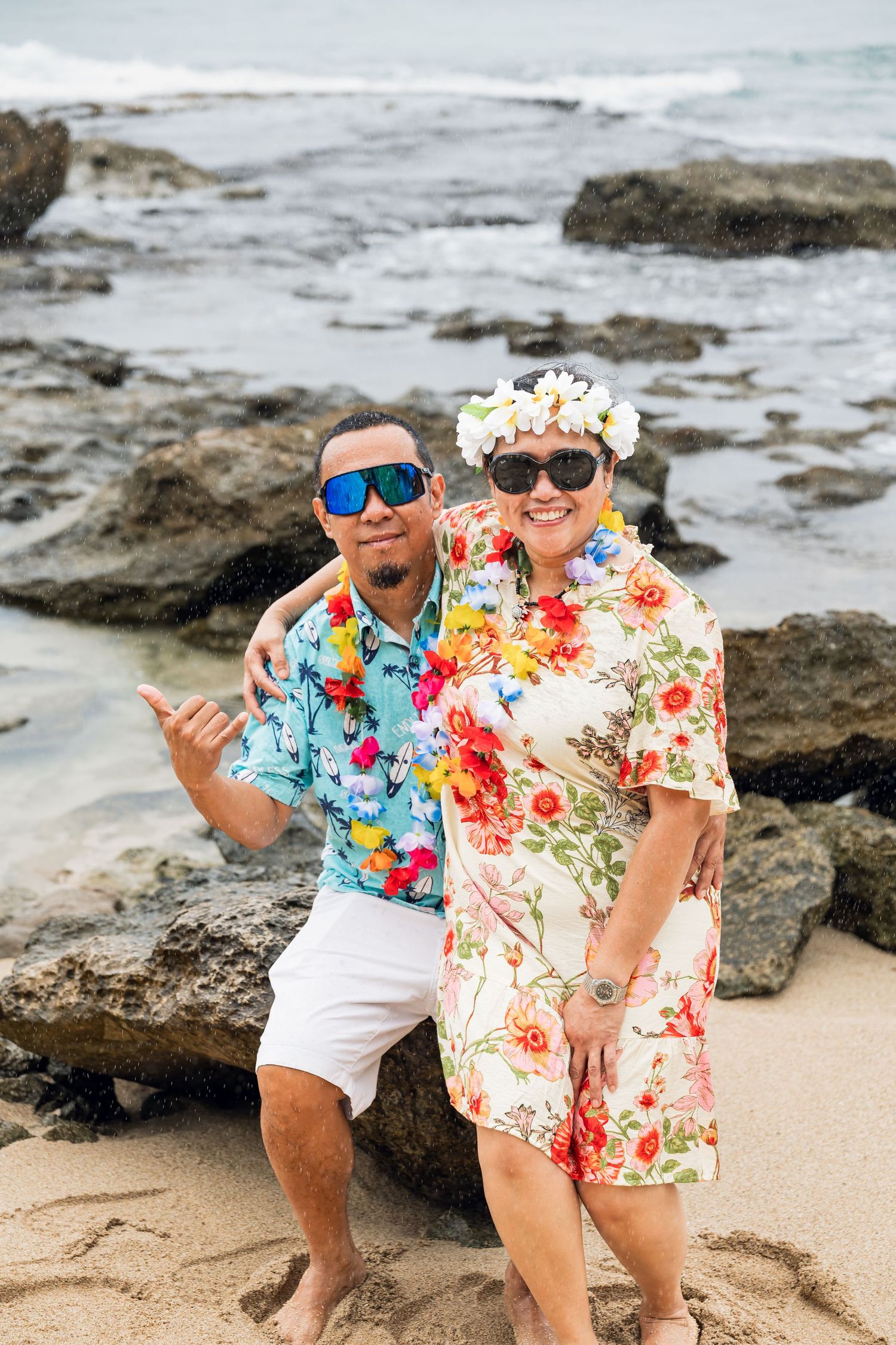 Couple in Hawaiian shirts and sunglasses posing on rocky beach, woman wearing floral lei and flower crown.