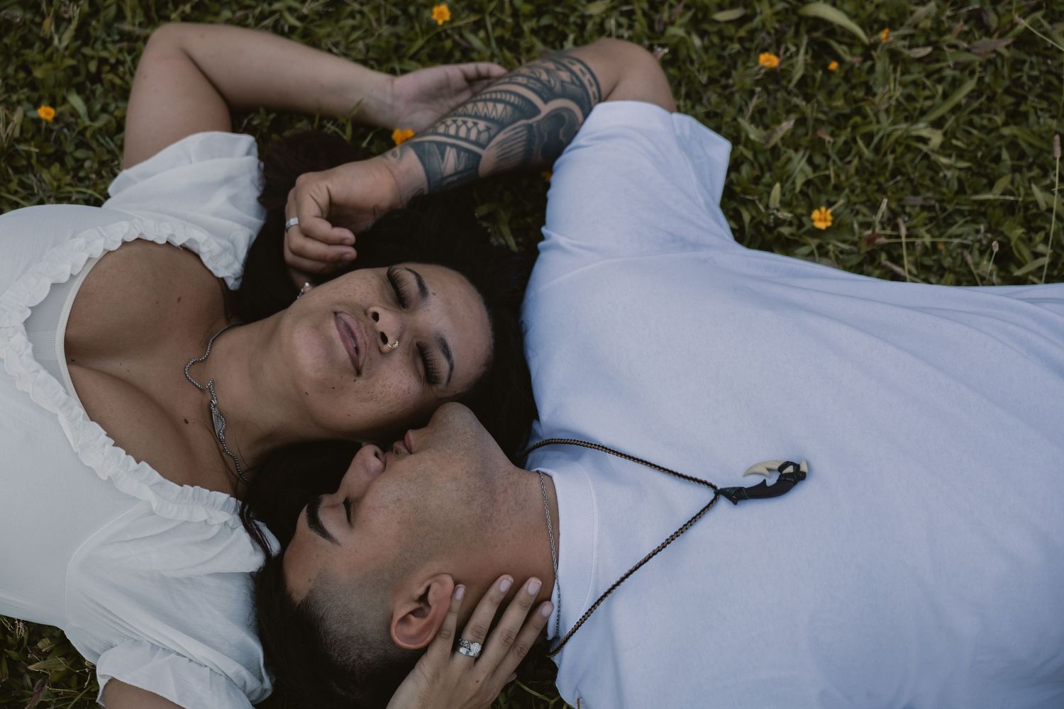 Couple lying on grass, woman with tattoos and man in white shirt sharing a tender moment.