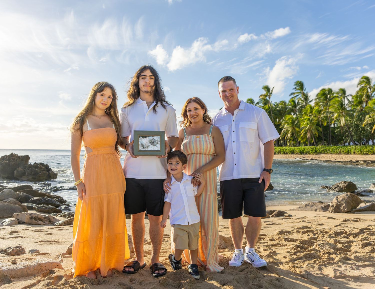 Family of five posing on a tropical beach with palm trees, holding a framed photo during a sunny vacation.