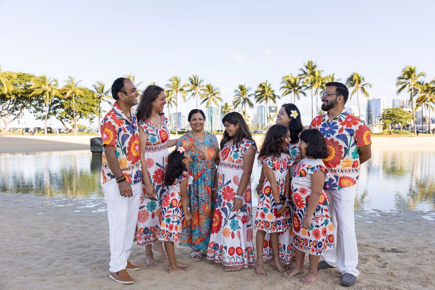 Family group photo on beach in matching colorful floral outfits with palm trees and city skyline backdrop.