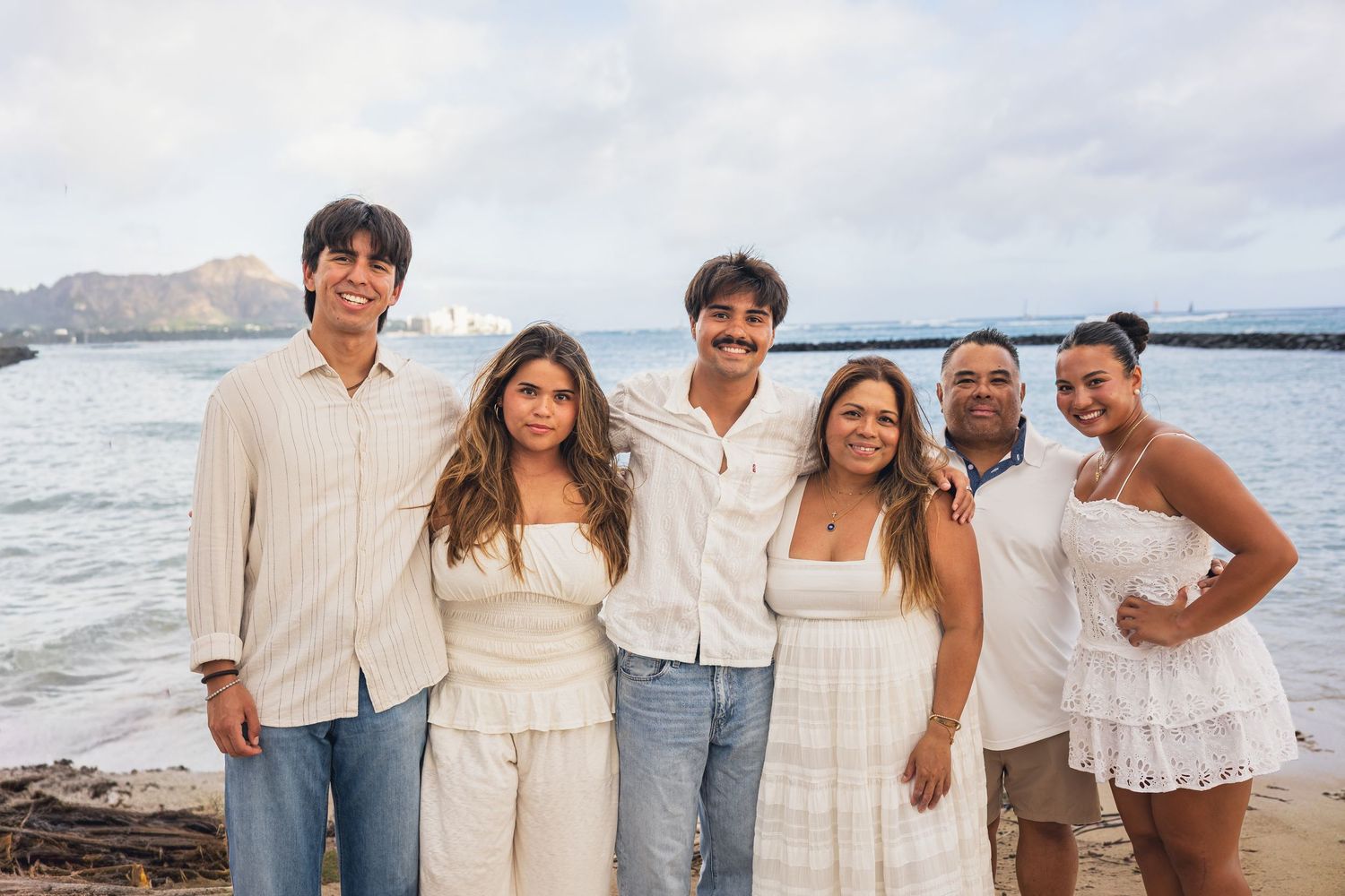 A family of six poses together on a beach wearing white and casual outfits with Diamond Head visible.