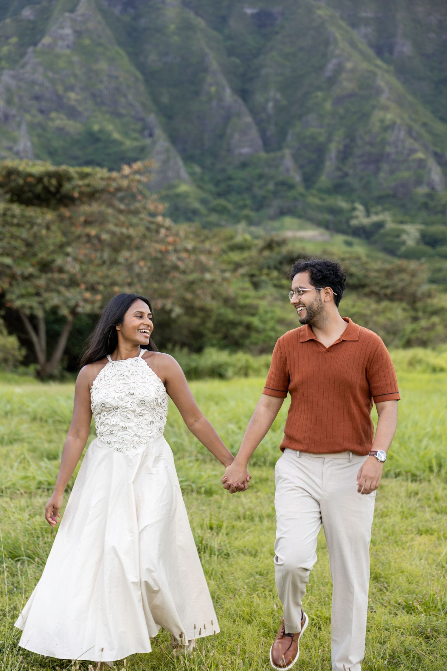 Happy couple holding hands walking through green meadow with stunning mountain range in Hawaii.