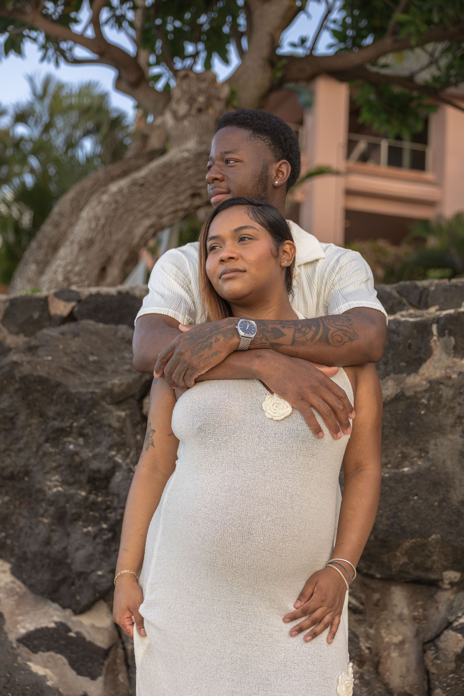 A pregnant couple poses outdoors, the man embracing the woman in a white dress near stone walls and tropical trees.