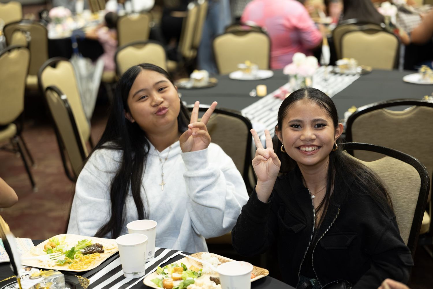 Two young women making peace signs while sitting at a banquet table with food at a formal event.