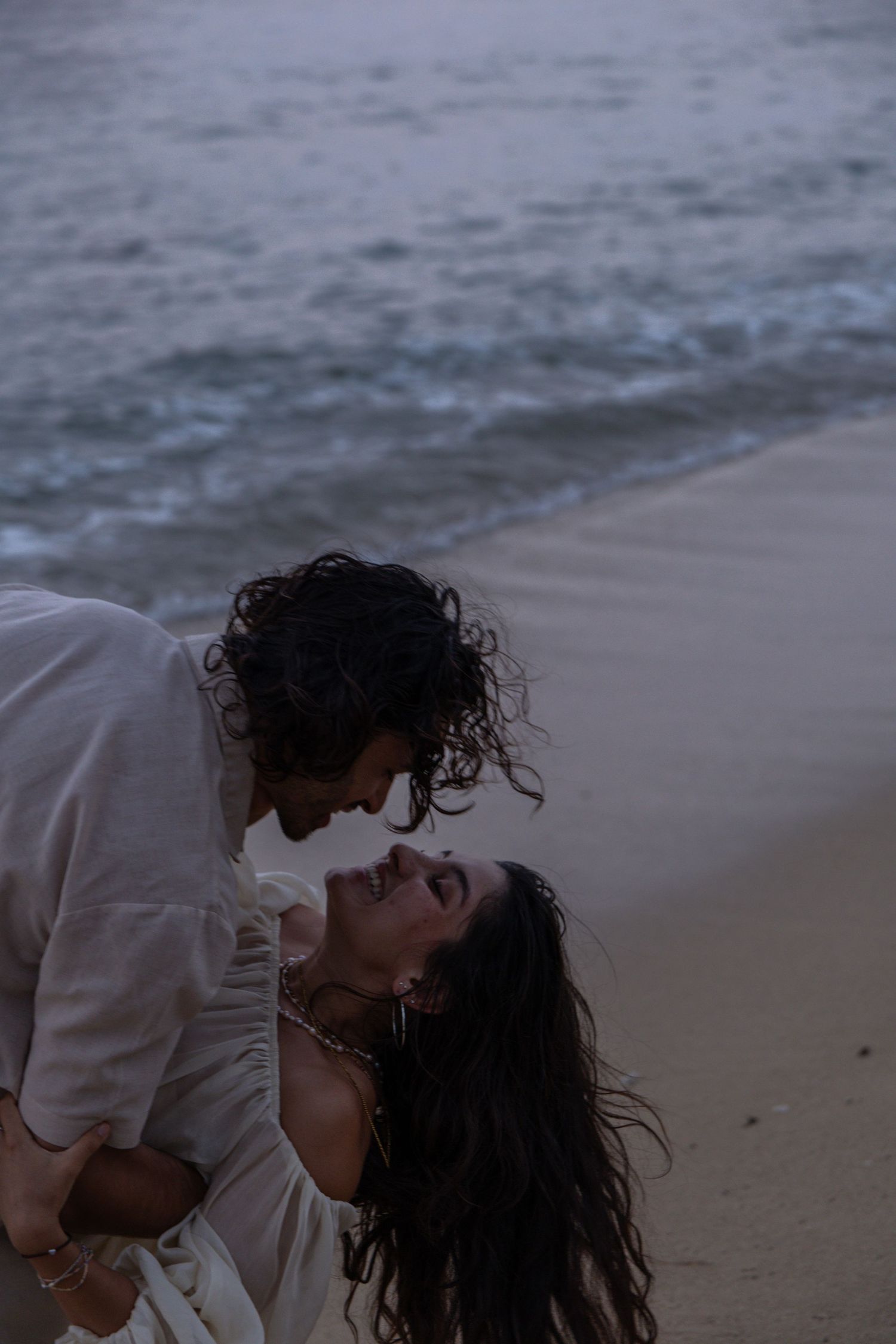 Romantic couple nearly kissing on a beach at dusk by ocean waves.