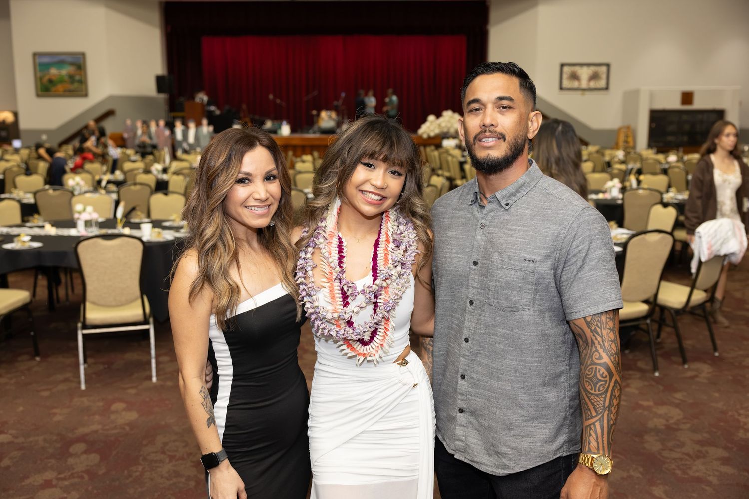 Three friends pose together at a formal banquet event, with the middle person wearing a floral lei.