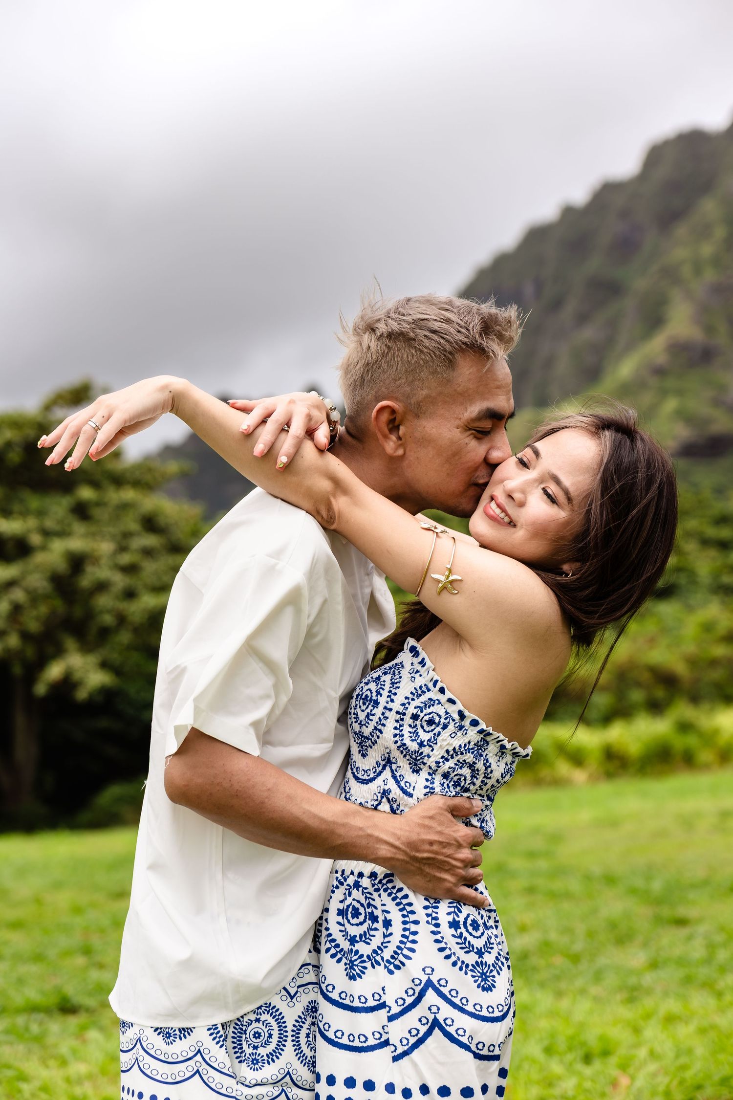 Man kisses smiling woman on cheek while embracing outdoors with lush green mountains behind them.