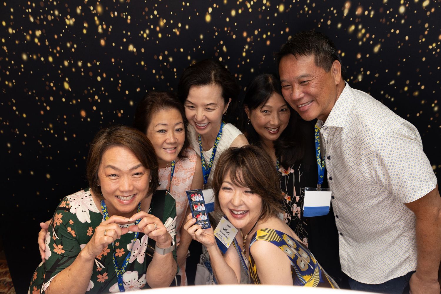 Group of six friends smiling and posing together in a photo booth with gold sparkle backdrop at an event.