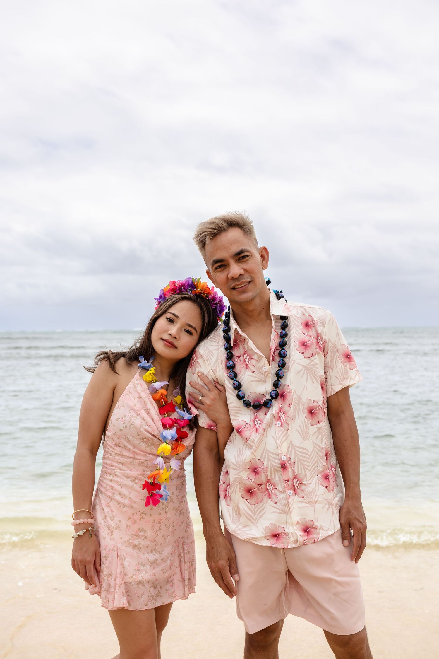 Couple in matching pink Hawaiian outfits with leis posing on a beach with ocean backdrop.