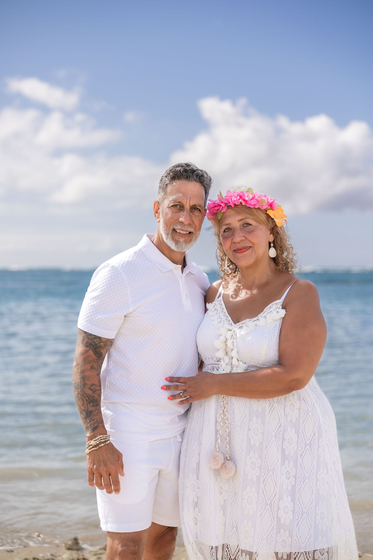 Mature couple in white outfits smile together on sunny Hawaiian beach, woman wearing pink flower crown.