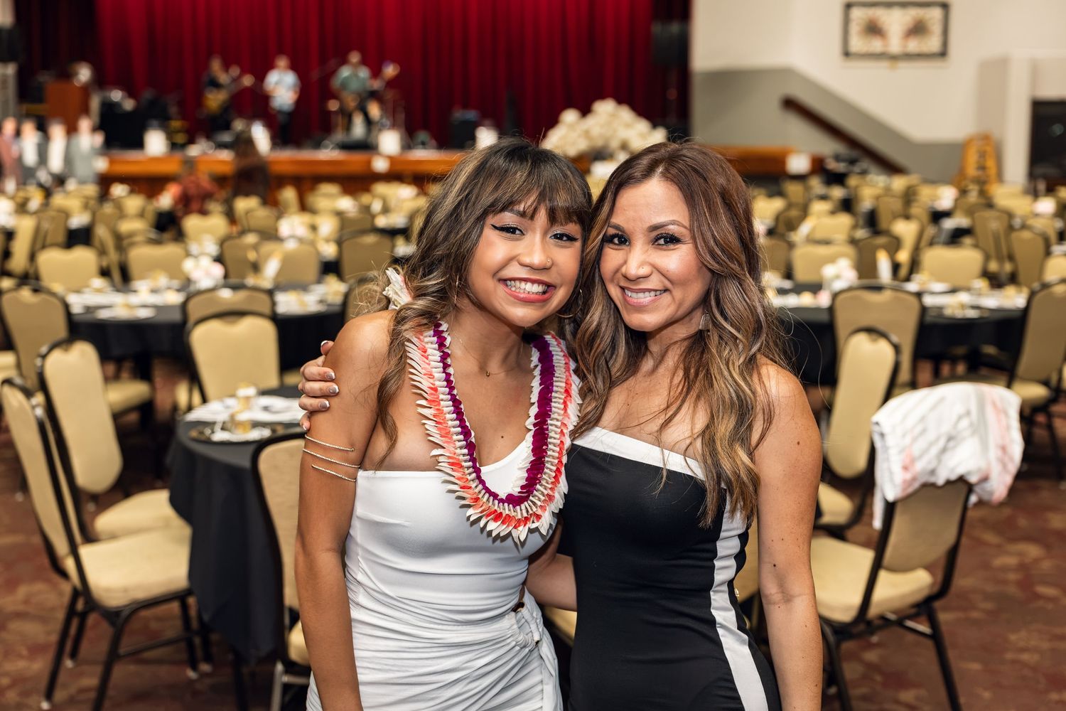Two smiling women posing together at an elegant banquet hall event, one wearing a floral lei.