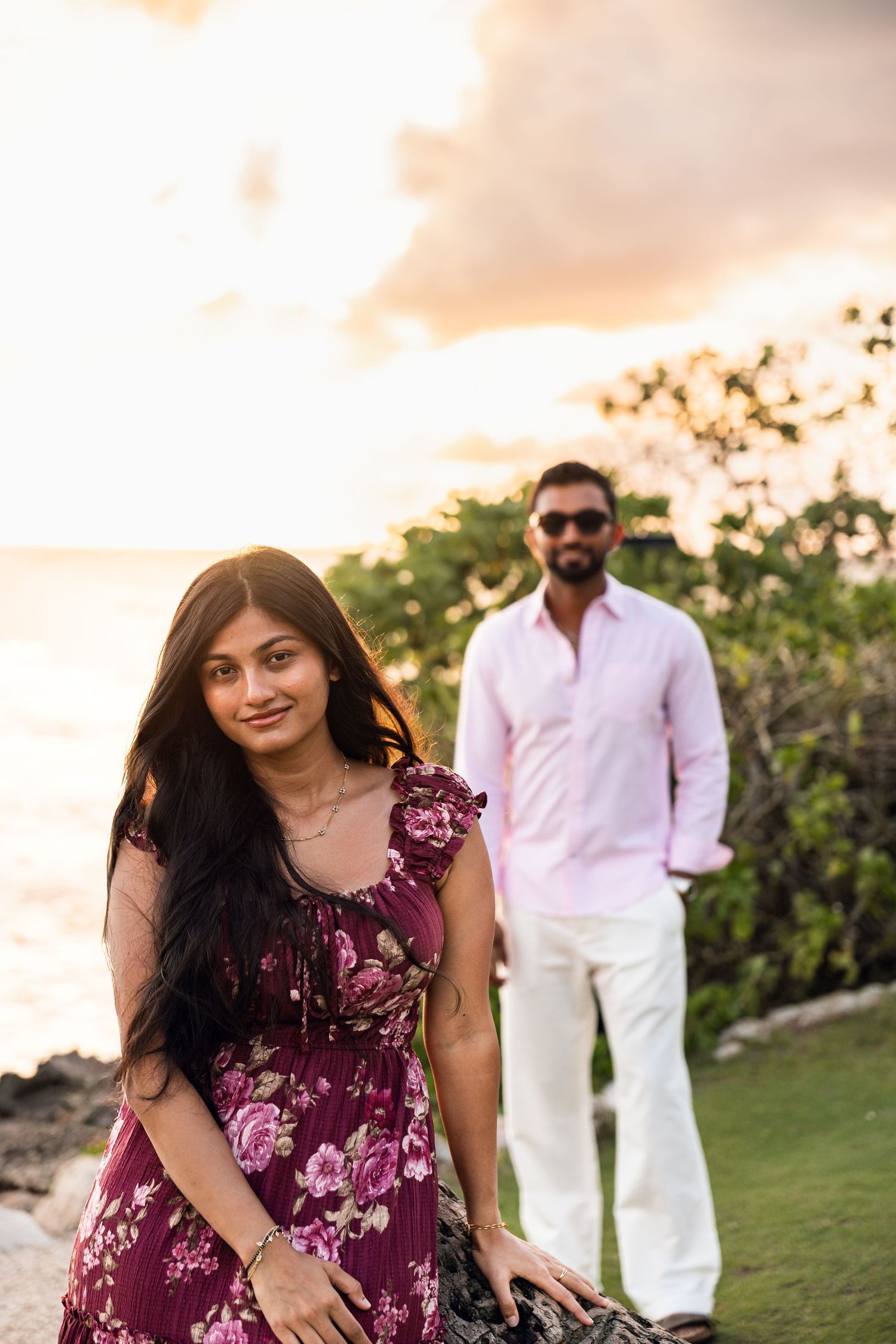 Woman in floral dress poses near ocean at sunset with man in pink shirt in background.