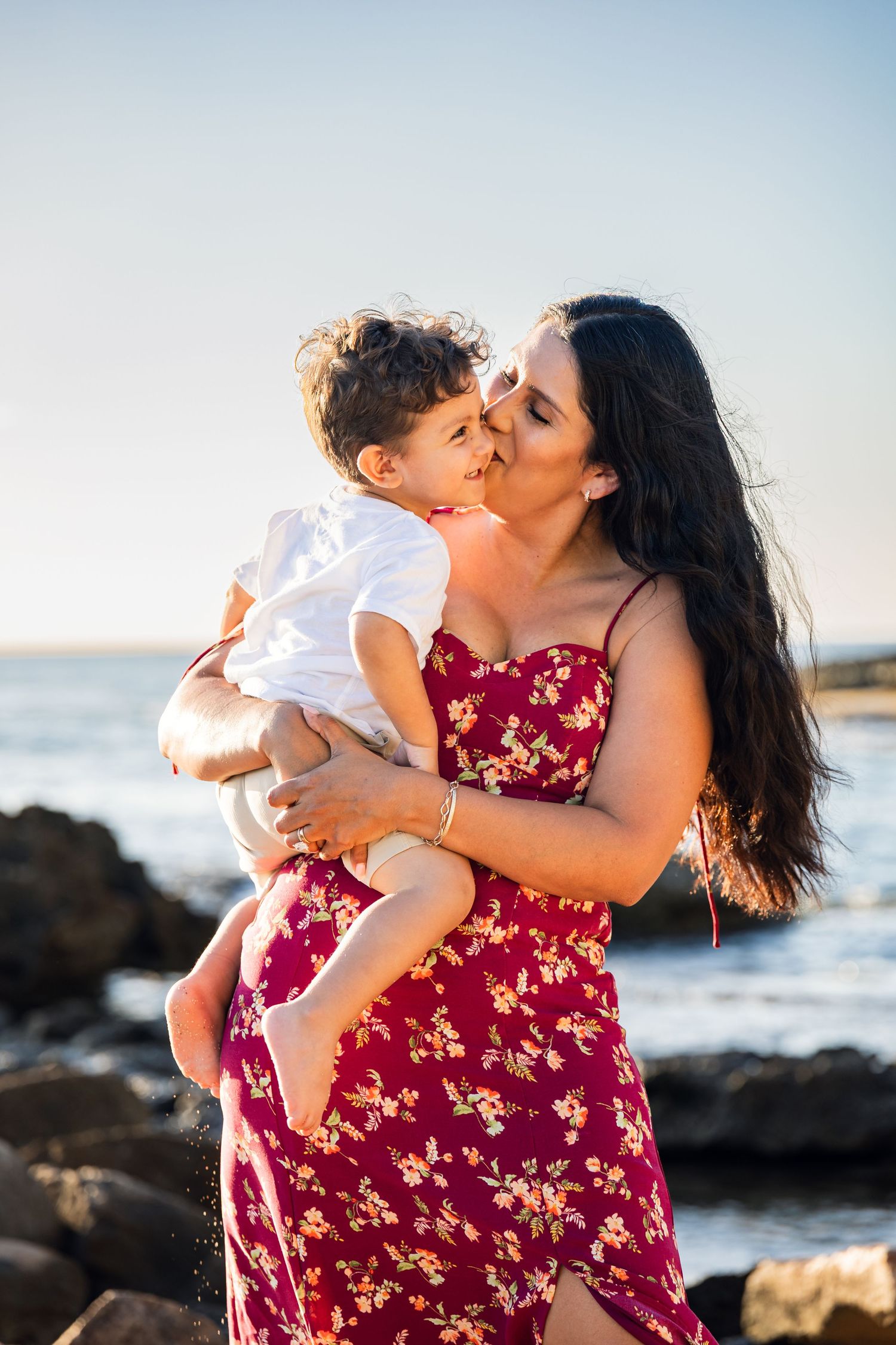 A mother in a red floral dress kisses her toddler son while holding him on a rocky beach at sunset.
