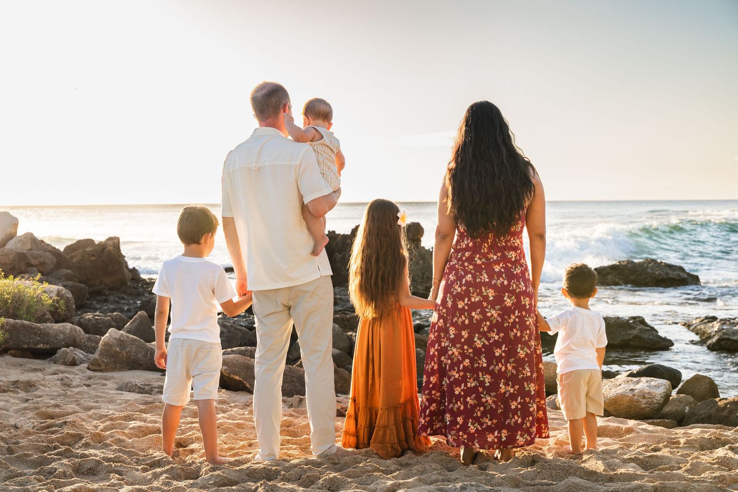 Family of five standing together on rocky beach watching sunset over ocean waves.