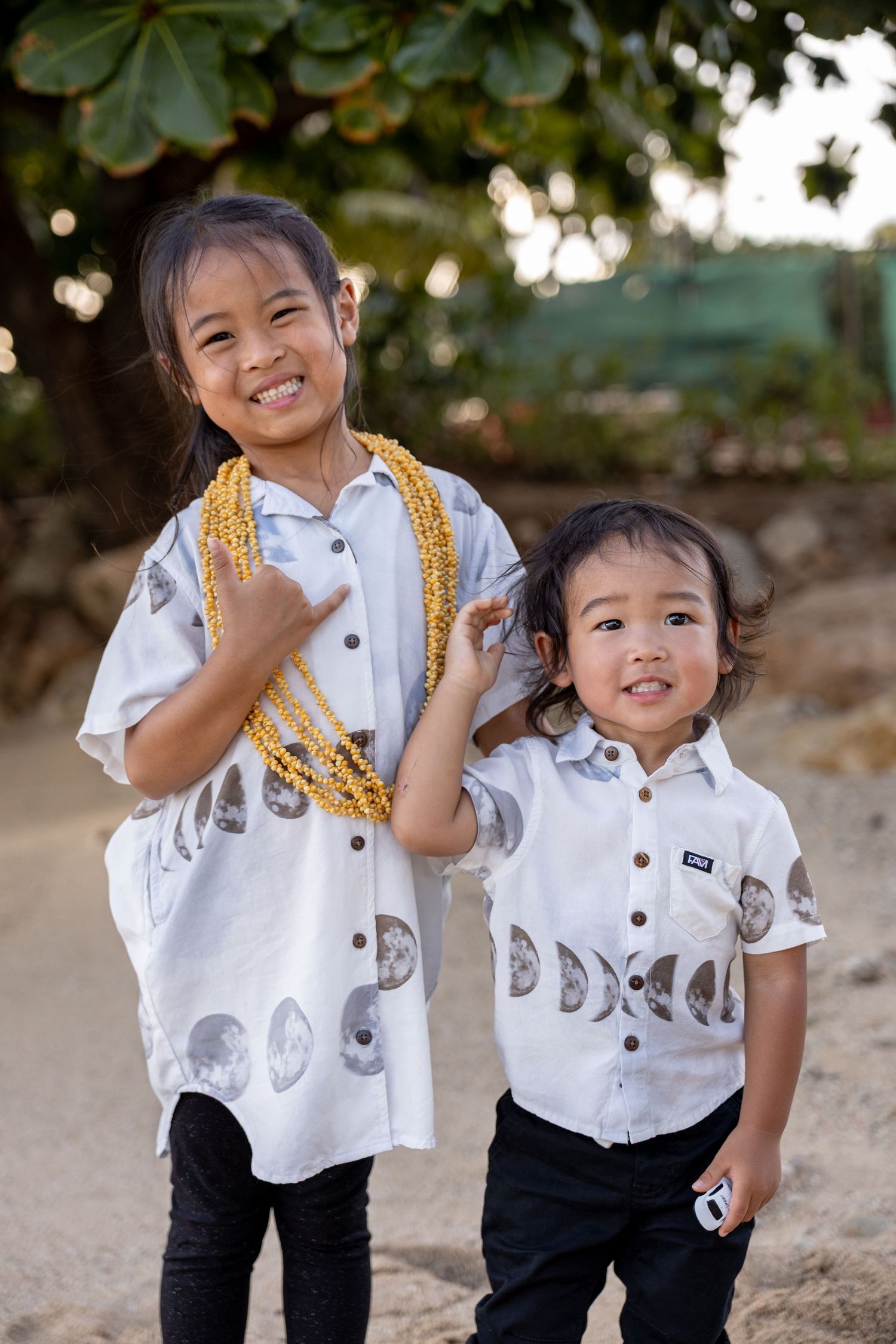 Two young siblings smile in matching moon-phase print white shirts outdoors near a tree.