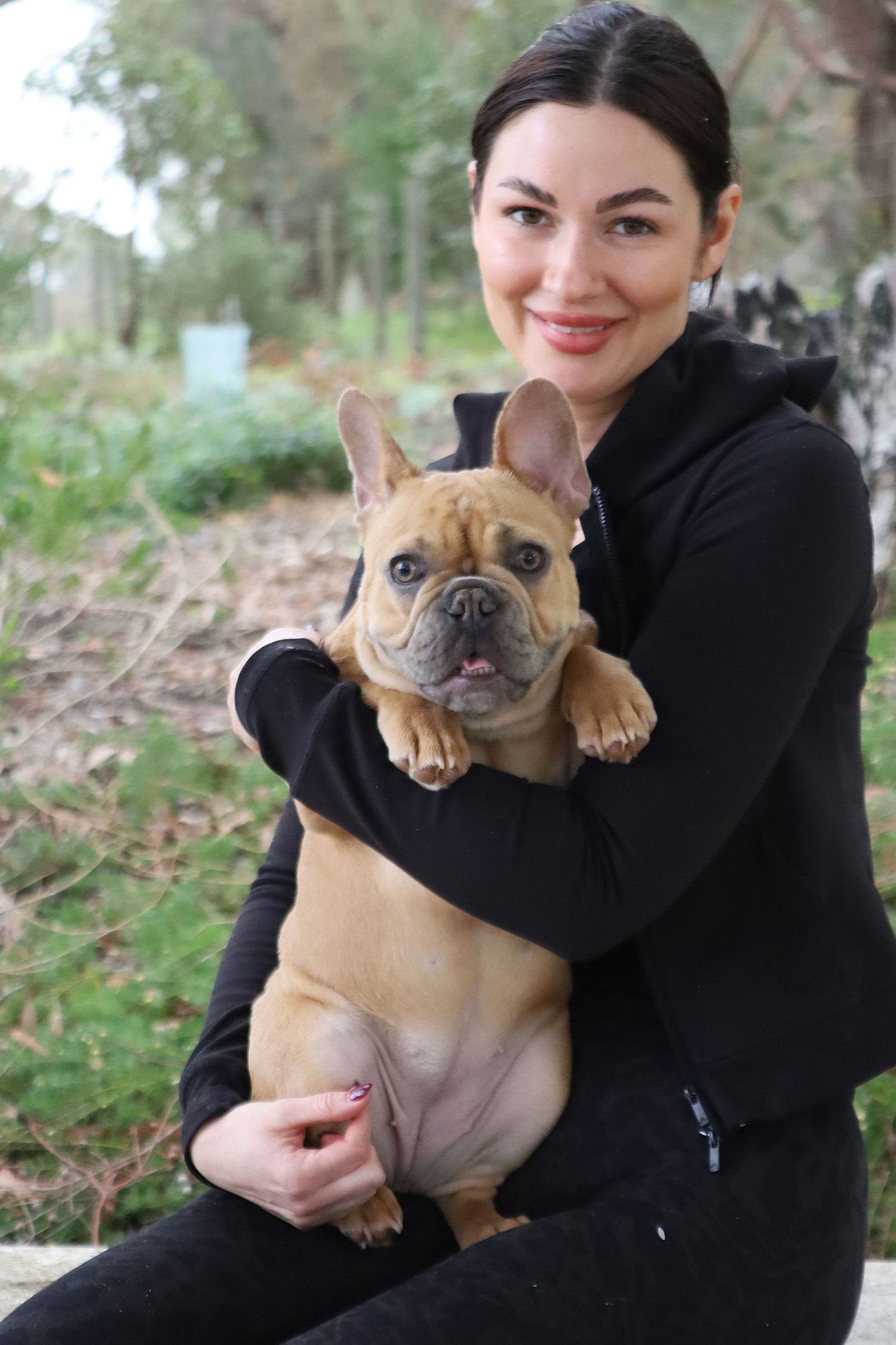 Attractive woman in black hoodie holding a fawn French Bulldog outdoors in a park.
By Chris Harban