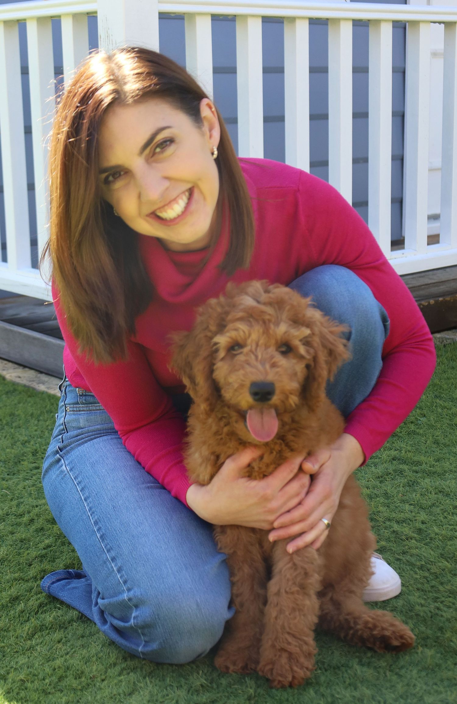Woman in pink top smiling while hugging an adorable fluffy golden doodle puppy outdoors on green grass.