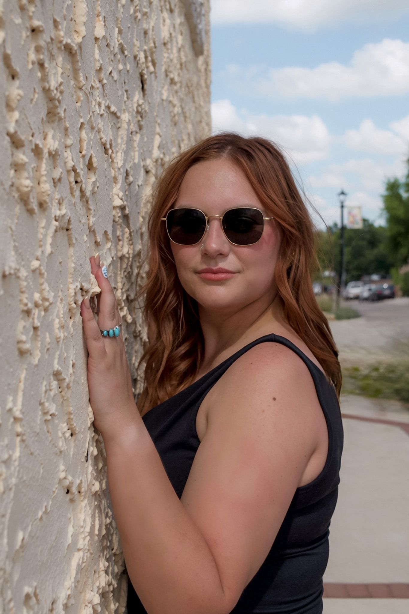 Young redhead woman in black dress wearing sunglasses leaning against textured stone wall outdoors.