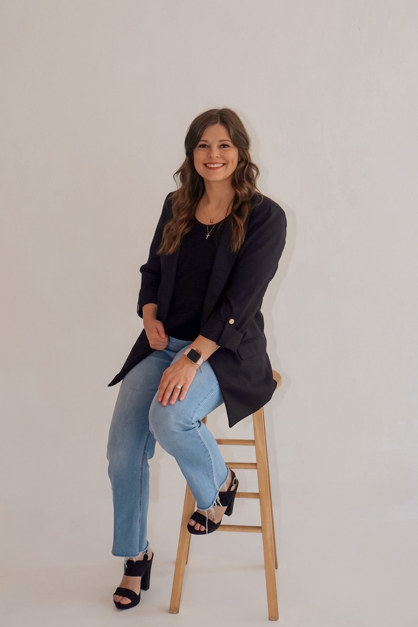 Smiling woman in black blazer and jeans sitting on wooden stool against white background.
