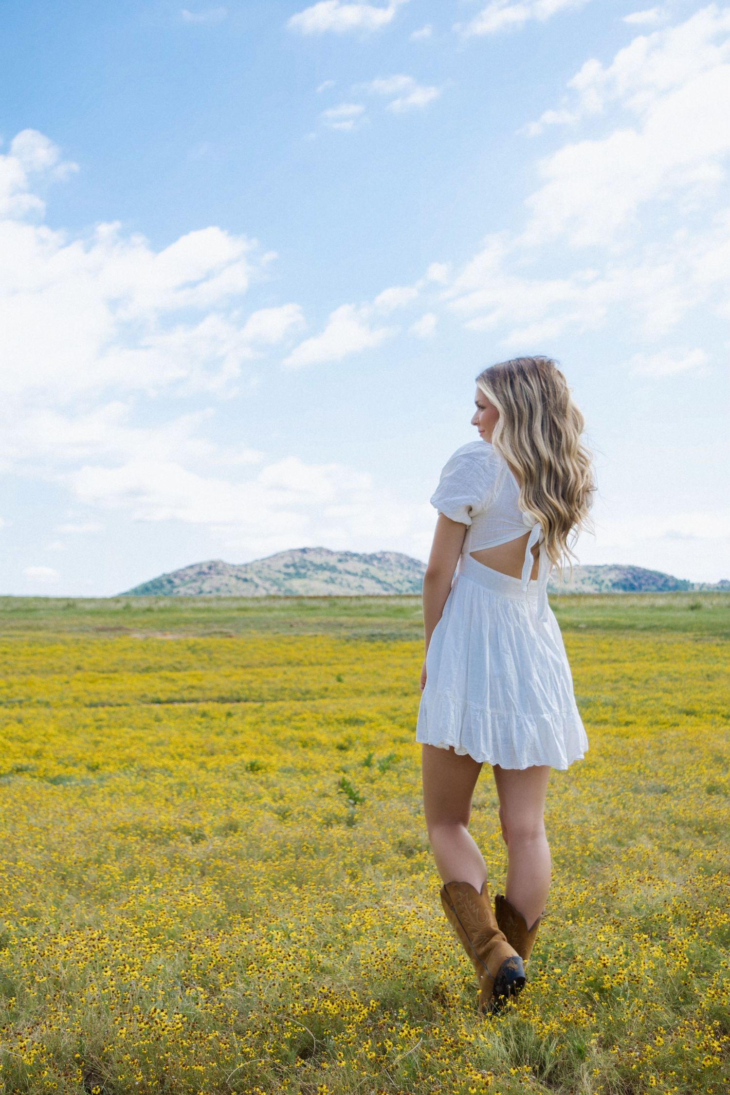 Young woman in white dress and cowboy boots standing in yellow wildflower field.