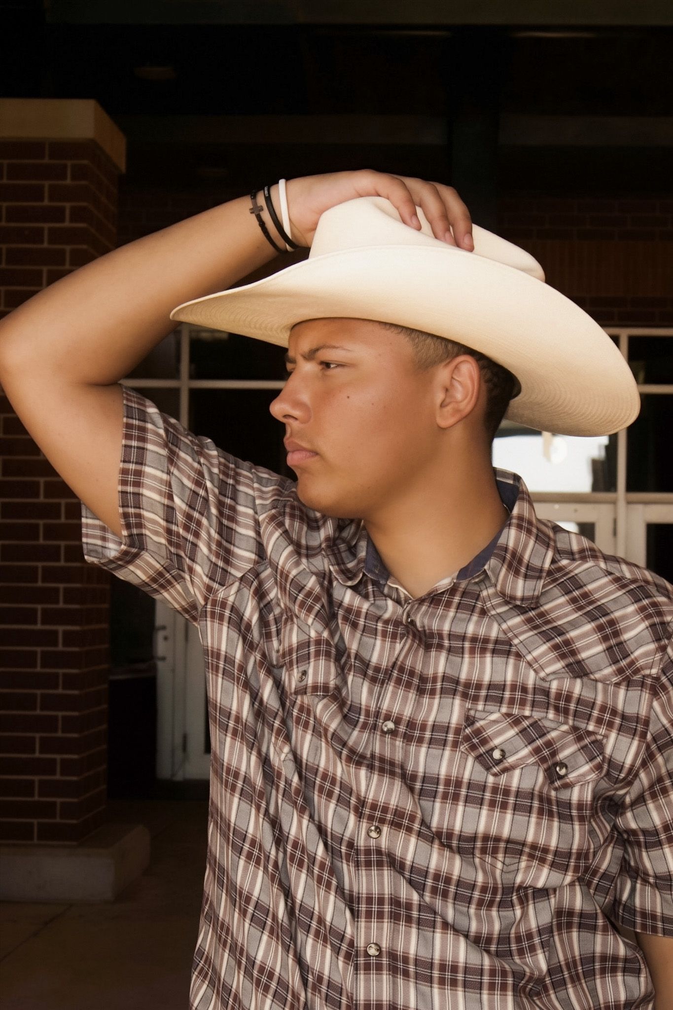 Young Hispanic man in plaid shirt tipping white cowboy hat outside brick building.