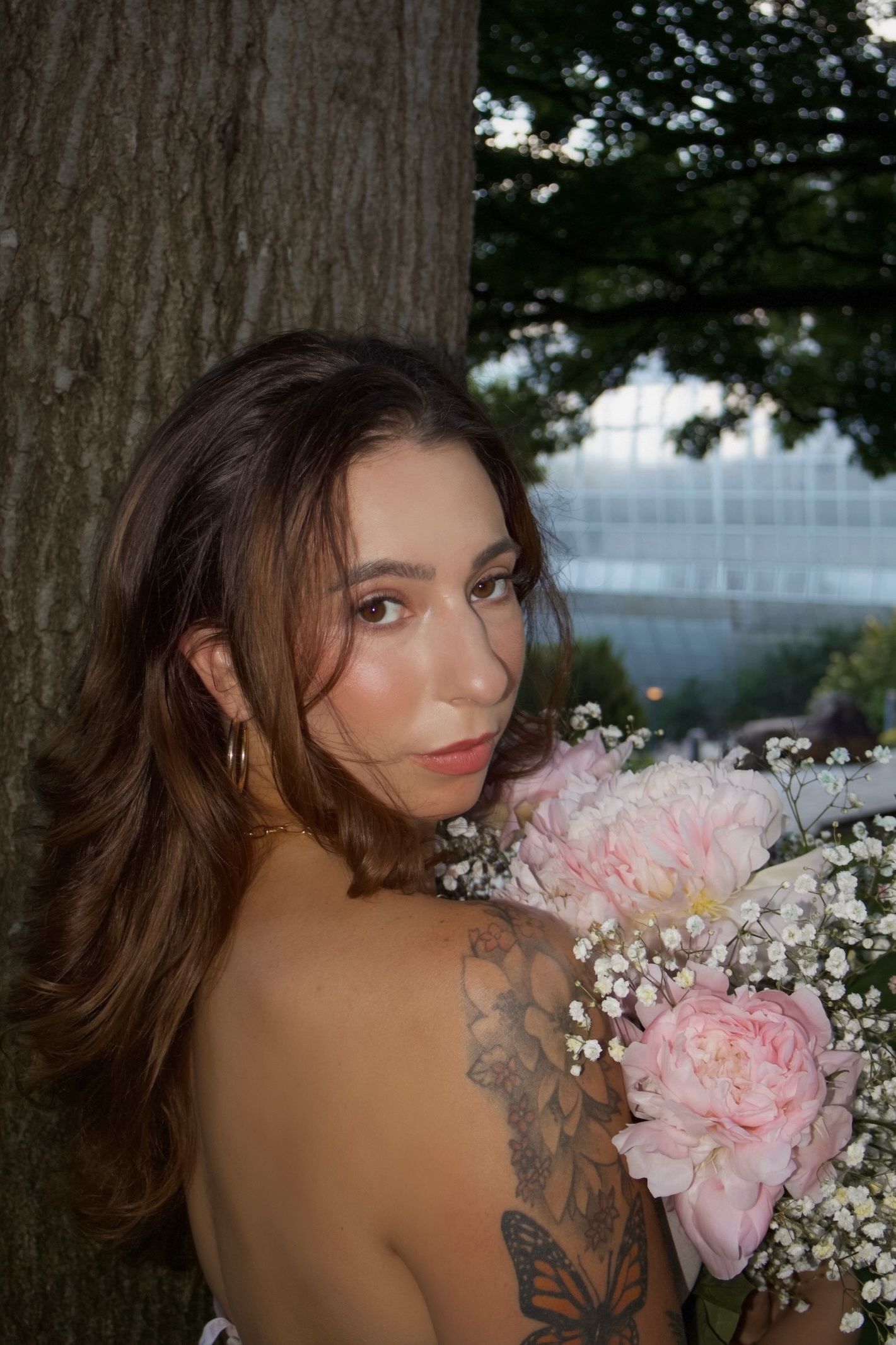 Young woman with butterfly tattoo holding pink peonies bouquet beside tree at dusk.