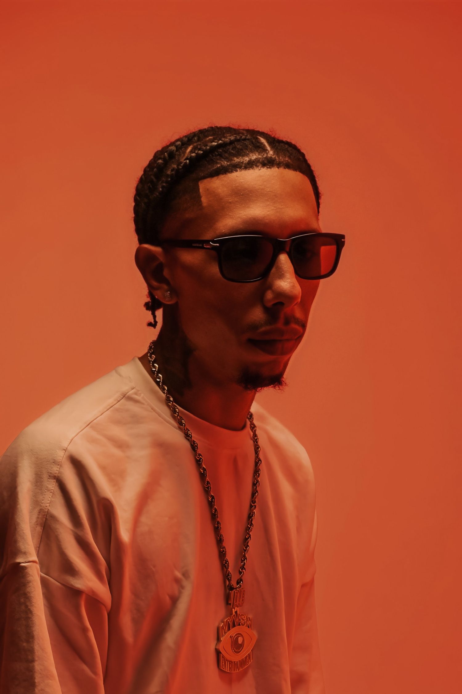 Stylish man with braids wearing sunglasses and gold chain necklace against orange-red background.
