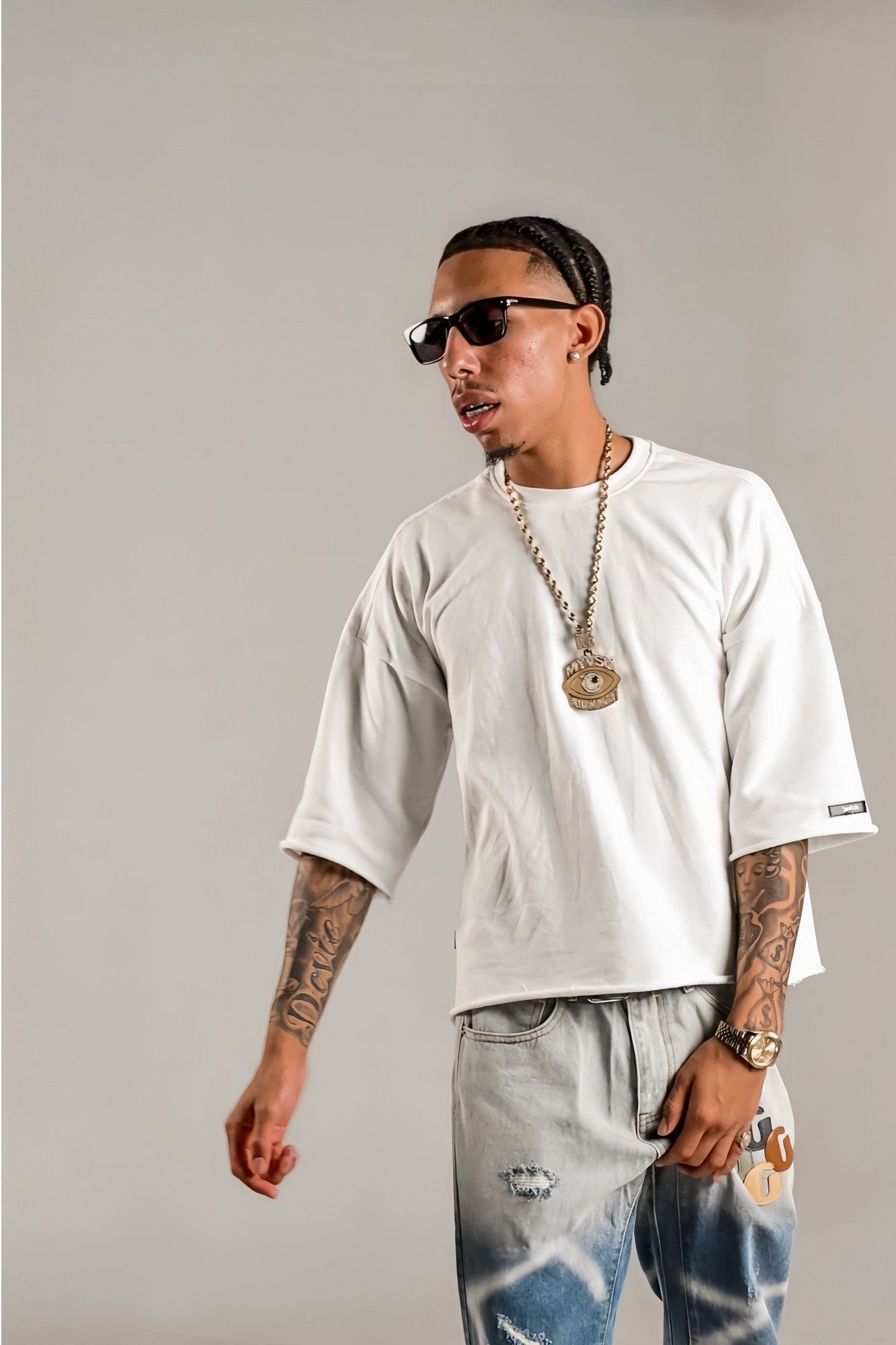 Tattooed man in white oversized tee, ripped jeans and gold chain necklace on gray background.