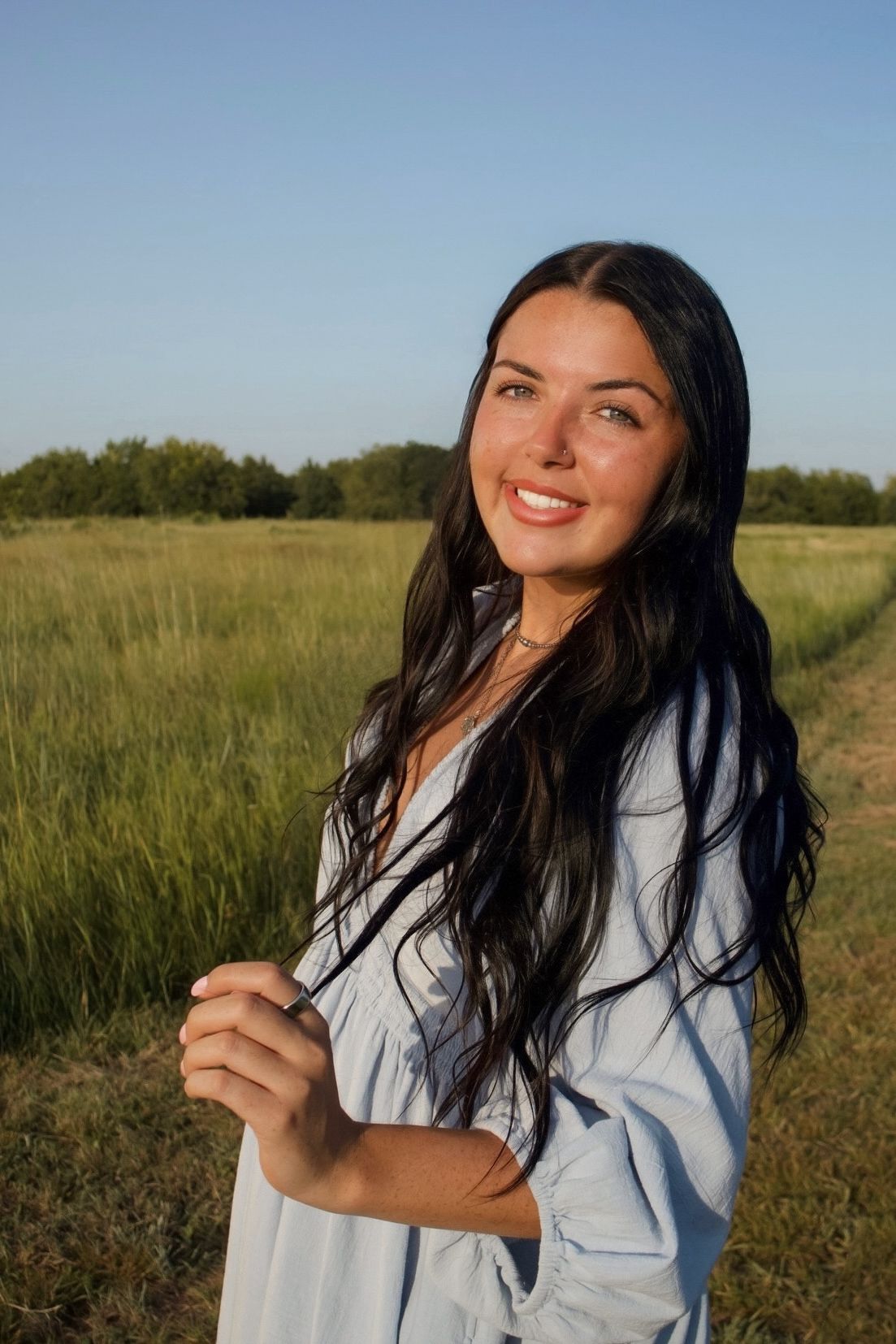 Smiling brunette woman in blue dress standing in sunny green field with clear blue sky.