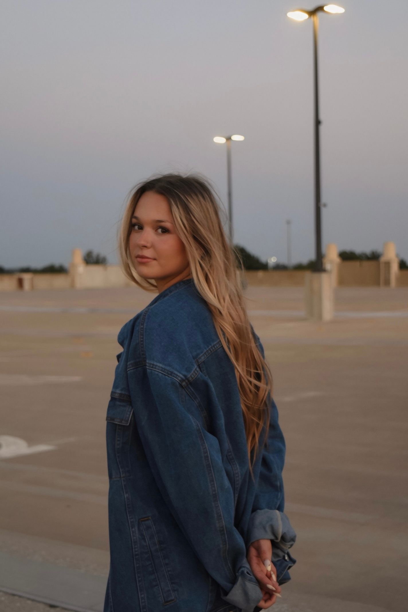 Blonde teen girl in oversized denim jacket looking back over shoulder in empty parking lot at sunset.