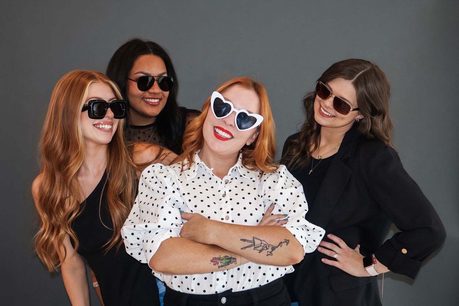 Four diverse women wearing stylish sunglasses pose together smiling against gray studio backdrop.