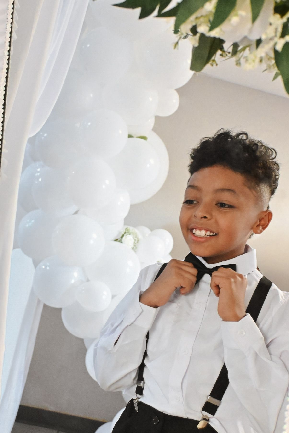 Smiling young ring bearer in white shirt suspenders and bow tie at wedding celebration.