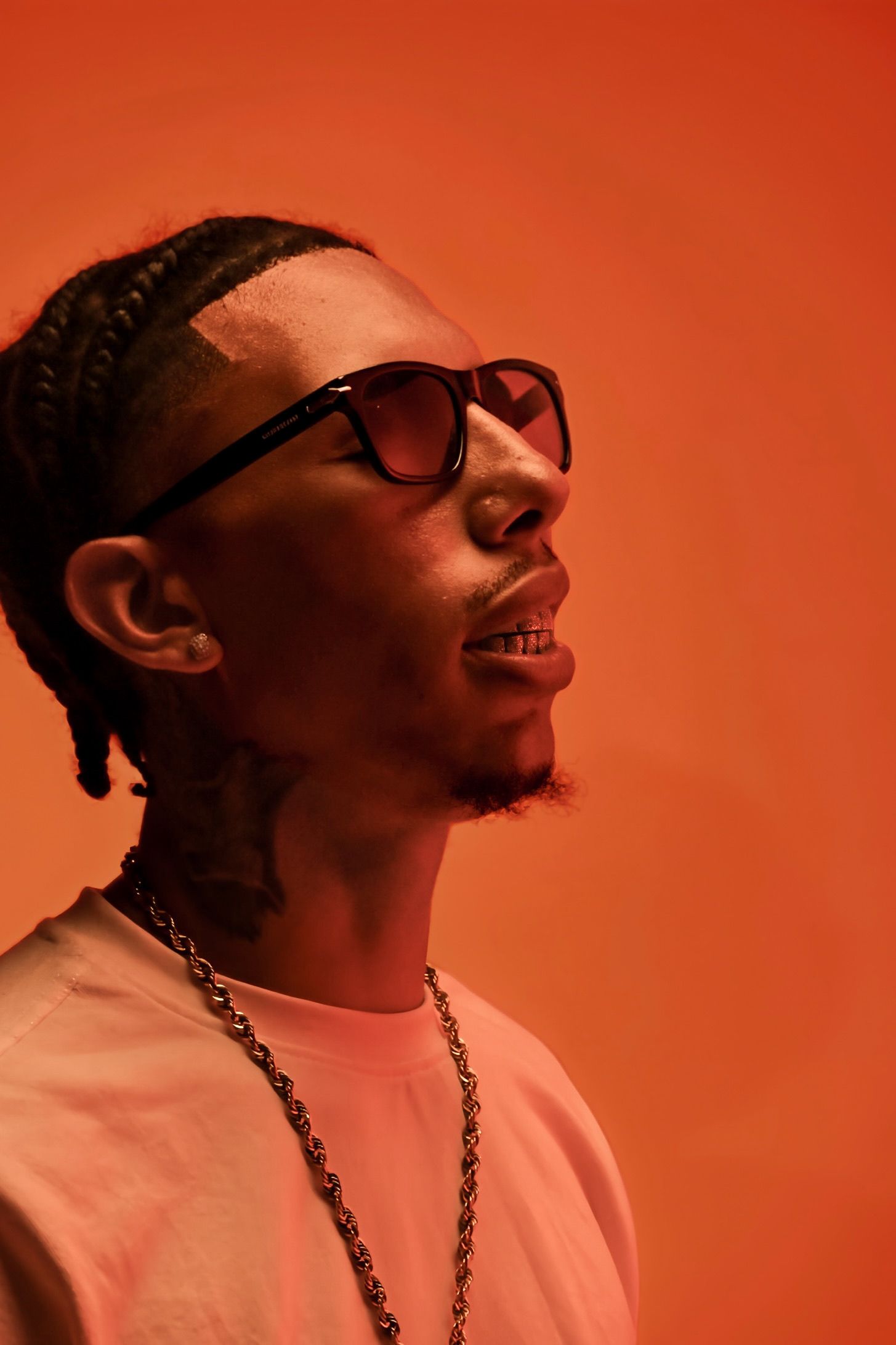 Close-up of man with braids and sunglasses smiling, wearing chain necklace on orange background.
