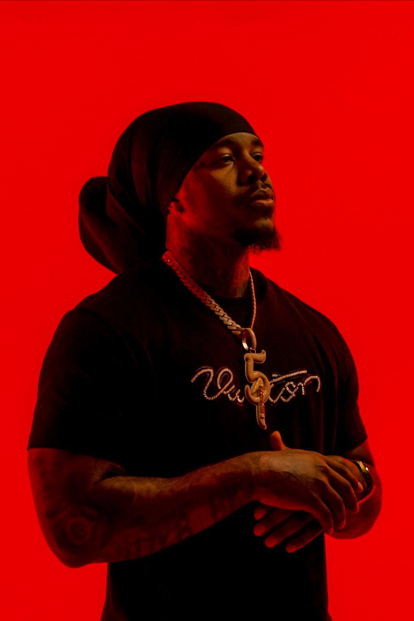 Man in black Louis Vuitton shirt with gold chain posing with arms crossed against red background.