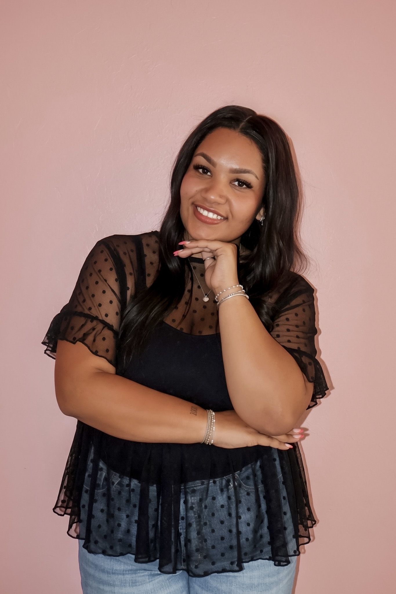 Smiling young woman in black polka dot sheer top posing with hand on chin against pink background.